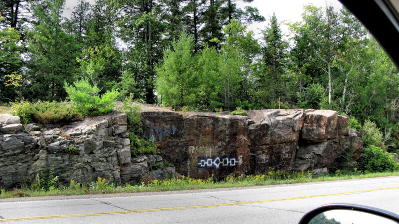 An image of the Hiawatha Wampum graffitied on a rock at the edge of a highway, taken from the driver's seat of a car, with a truck visible in the car's rear view mirror.